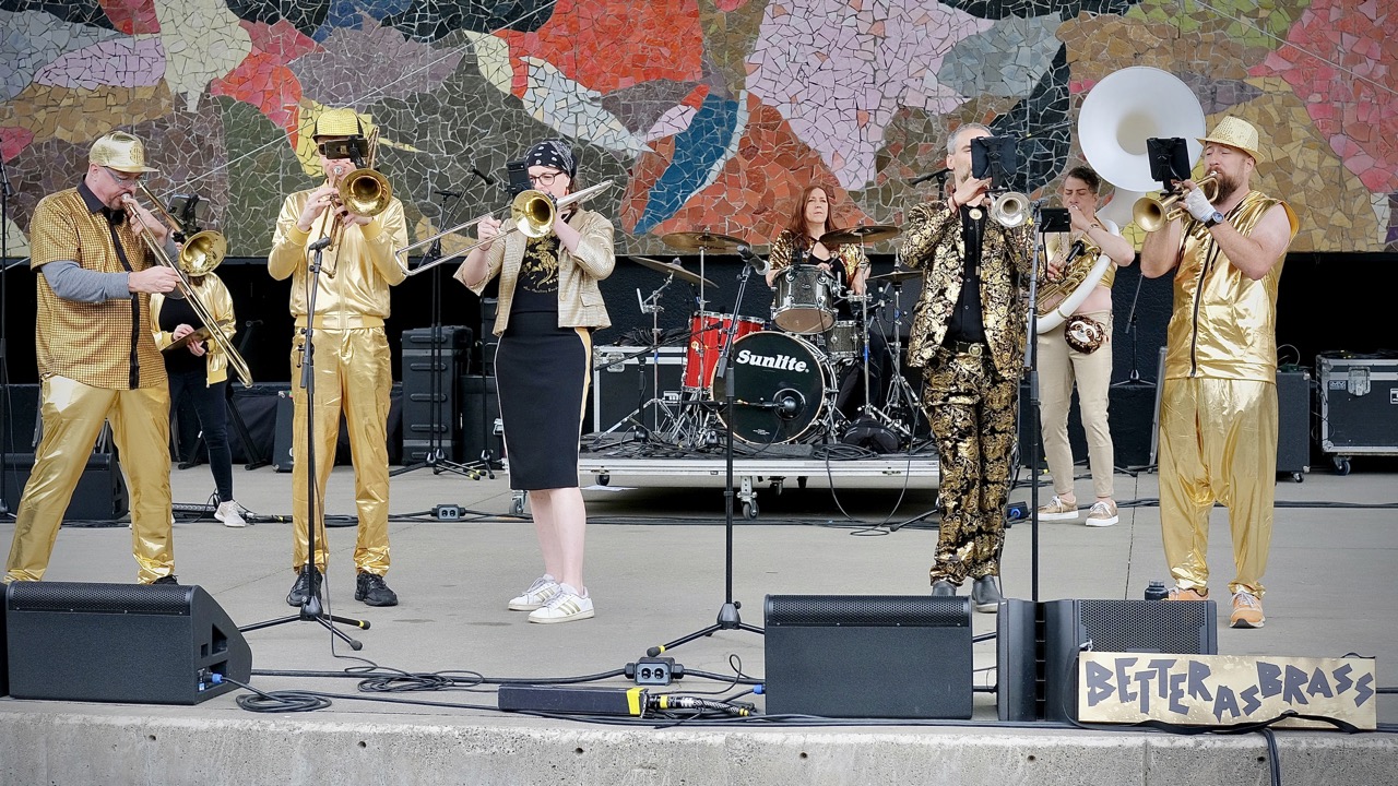 People on stage playing brass instruments and drums mostly dressed in metallic gold.